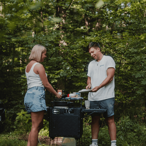 Ein Mann und eine Frau mit kurzen Hosen bei der Essenszubereitung mit einer Outdoorküche