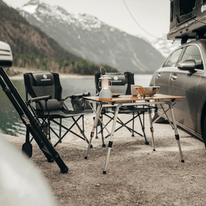 Nahaufnahme eines Campingplatzes mit einem Tisch, Stühlen und einer Lampe. Im Hintergrund befindet sich ein Gebirgszug. 